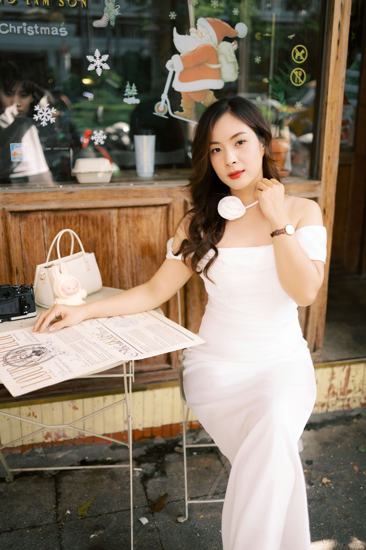 Woman in white dress sits at outdoor cafe table with Christmas decorations in window during daytime.