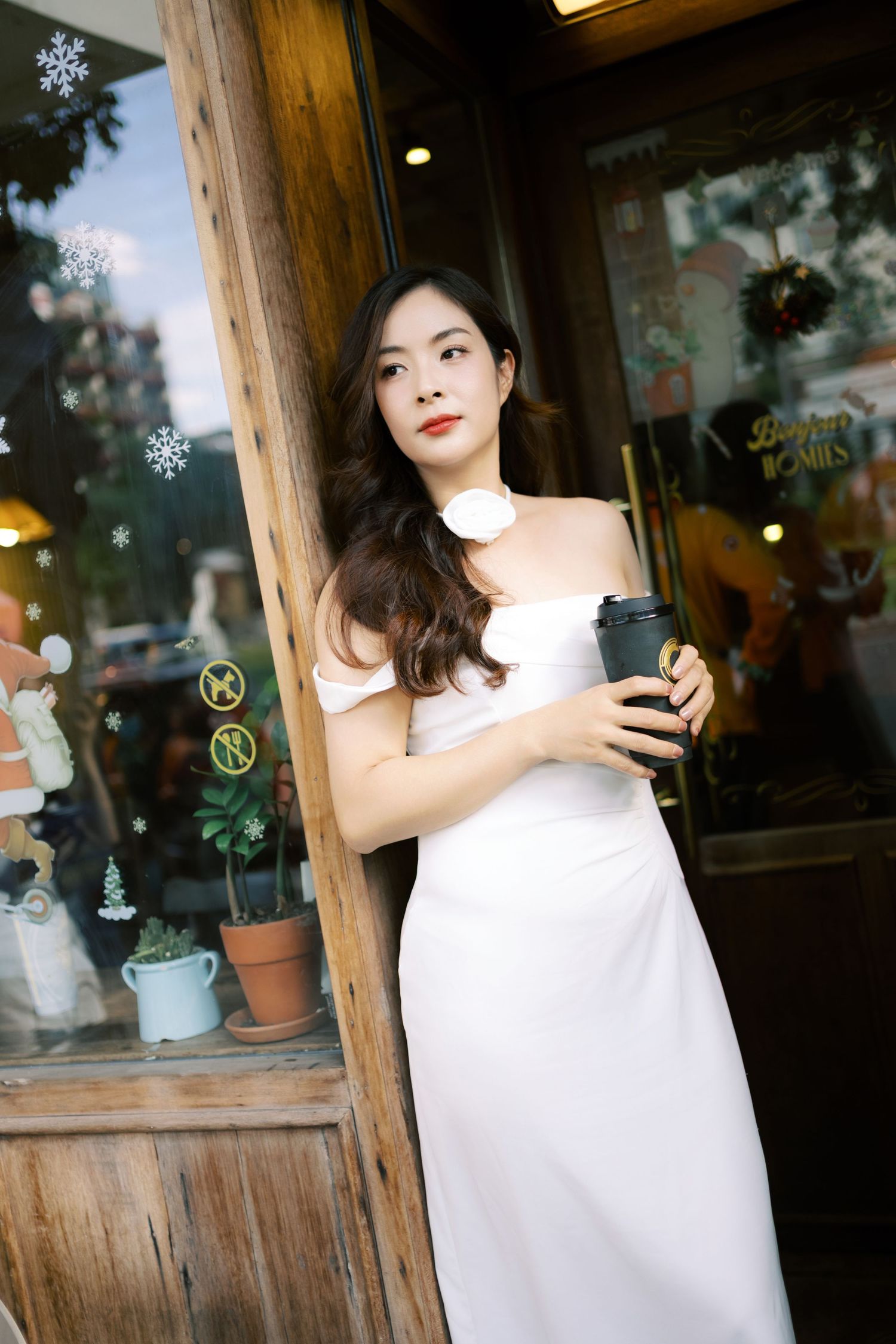 Person in white dress holds coffee cup while standing by rustic wooden storefront with snowflake decorations.