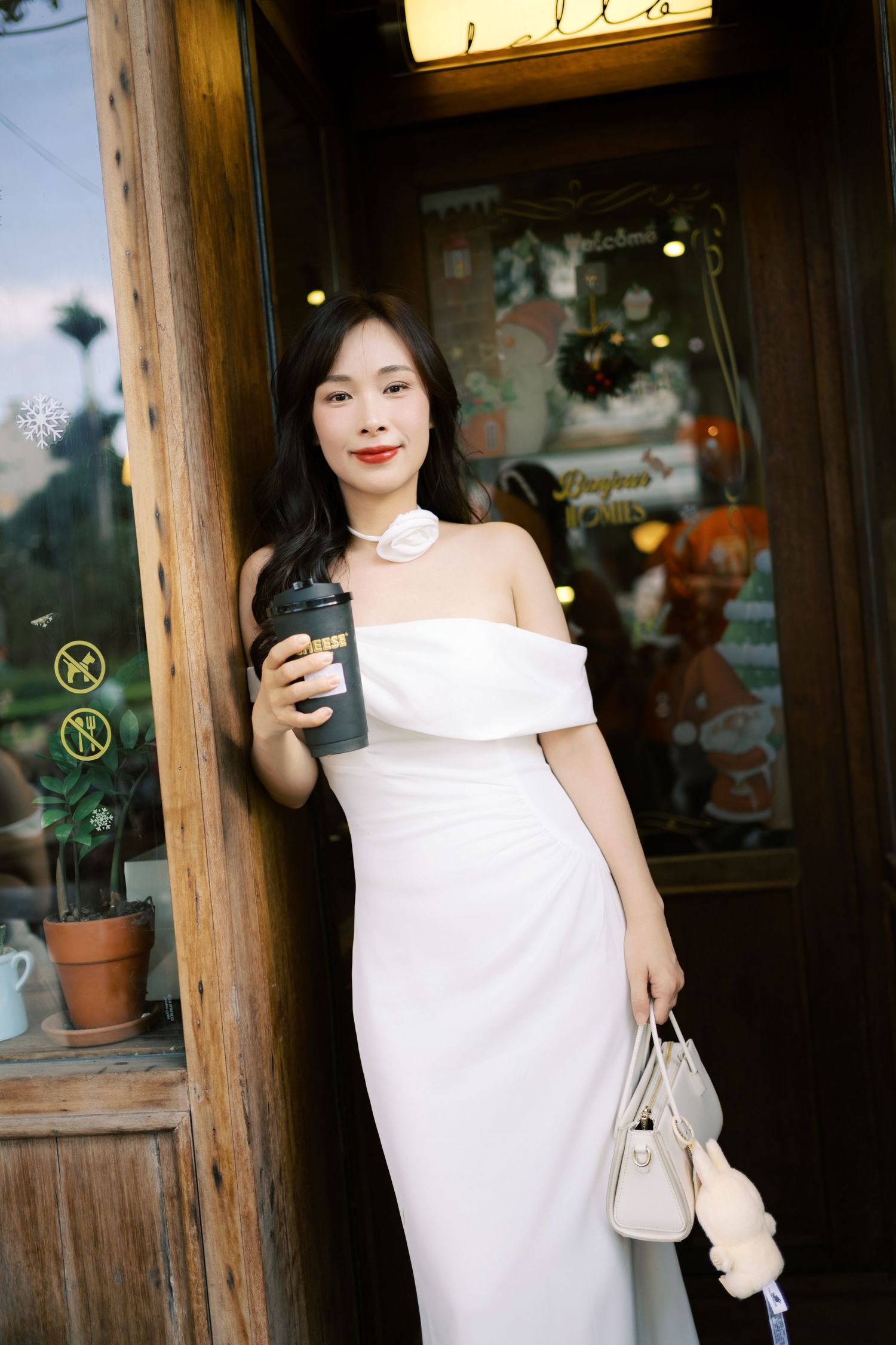 Someone in an elegant white off-shoulder dress stands in a doorway holding a coffee cup and small handbag.