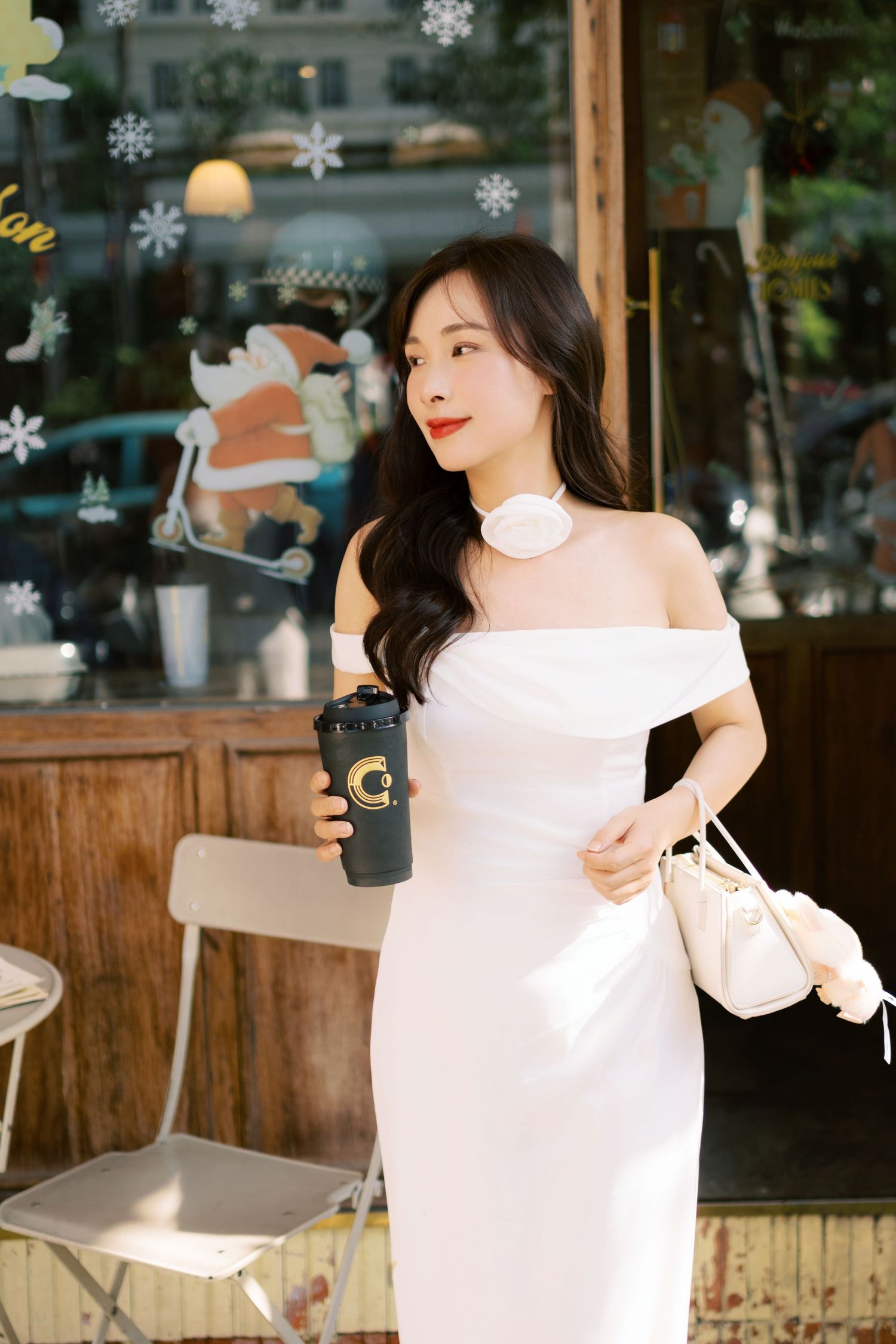 A person in a white dress holding a coffee cup stands at a cafe counter with snowflake decorations on the window.