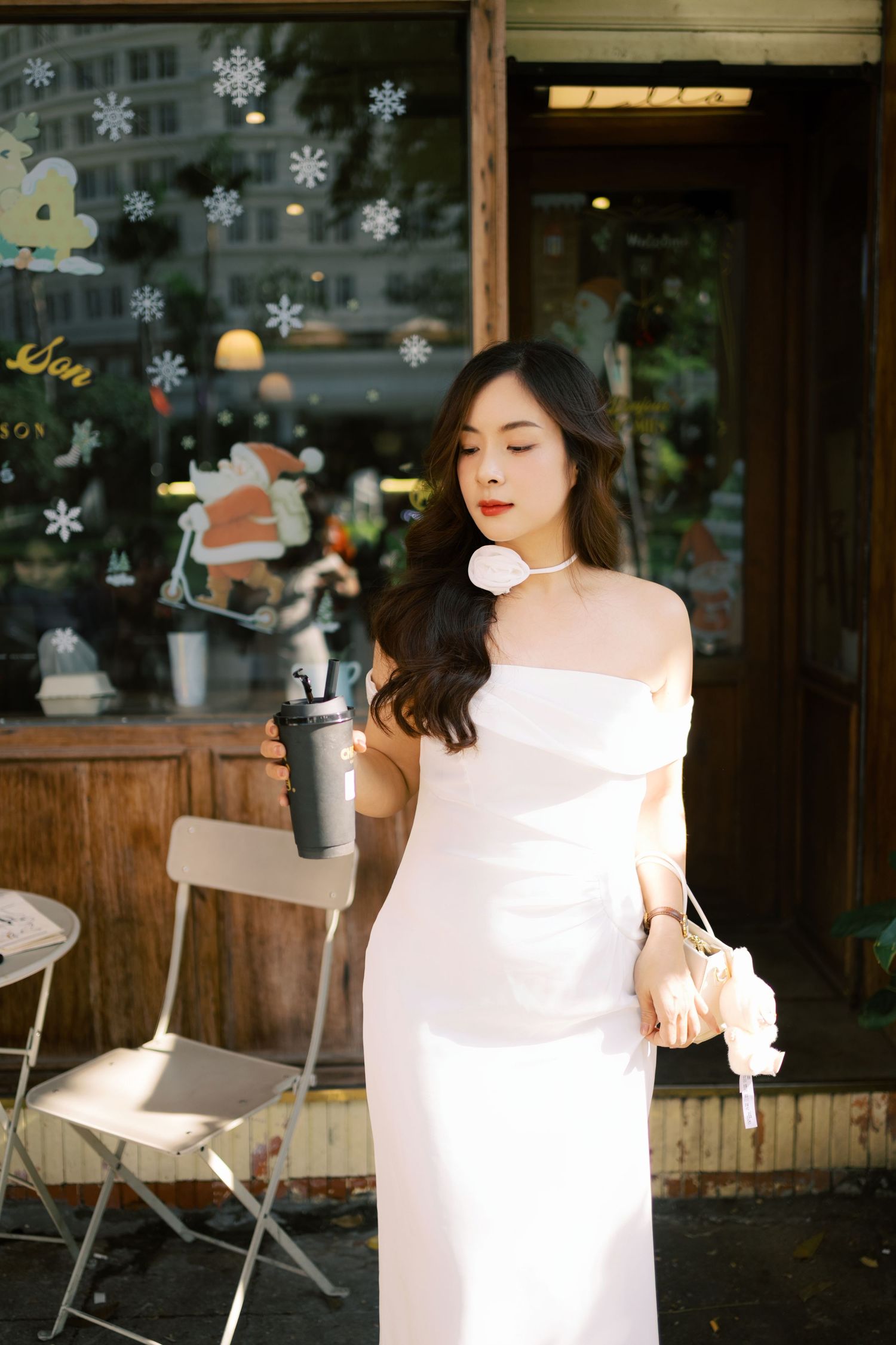 Elegant off-shoulder white dress in front of cafe window decorated with snowflakes and holiday decor.