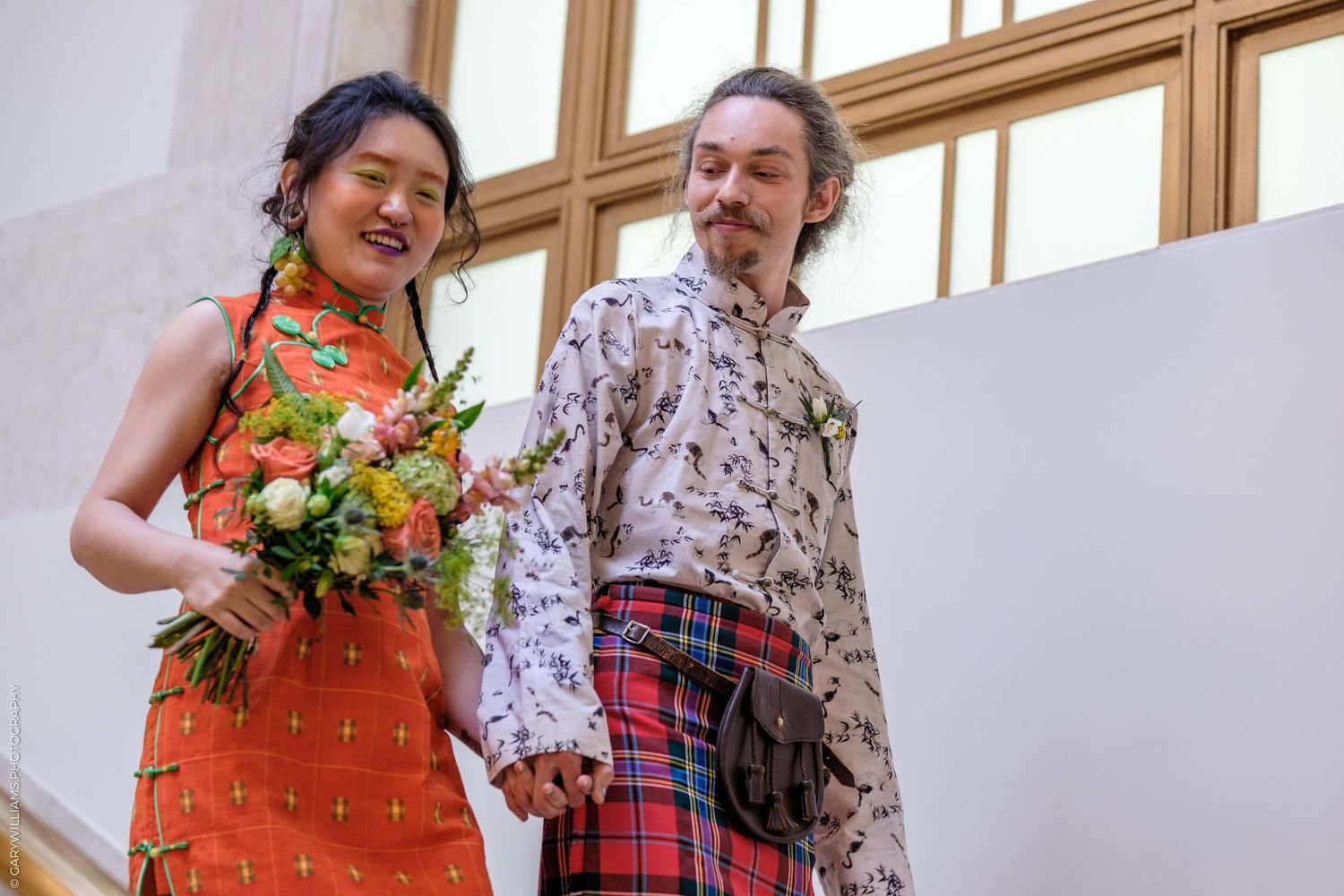 A couple wearing a bright orange dress and floral pattern shirt pose together in a sunlit interior space.