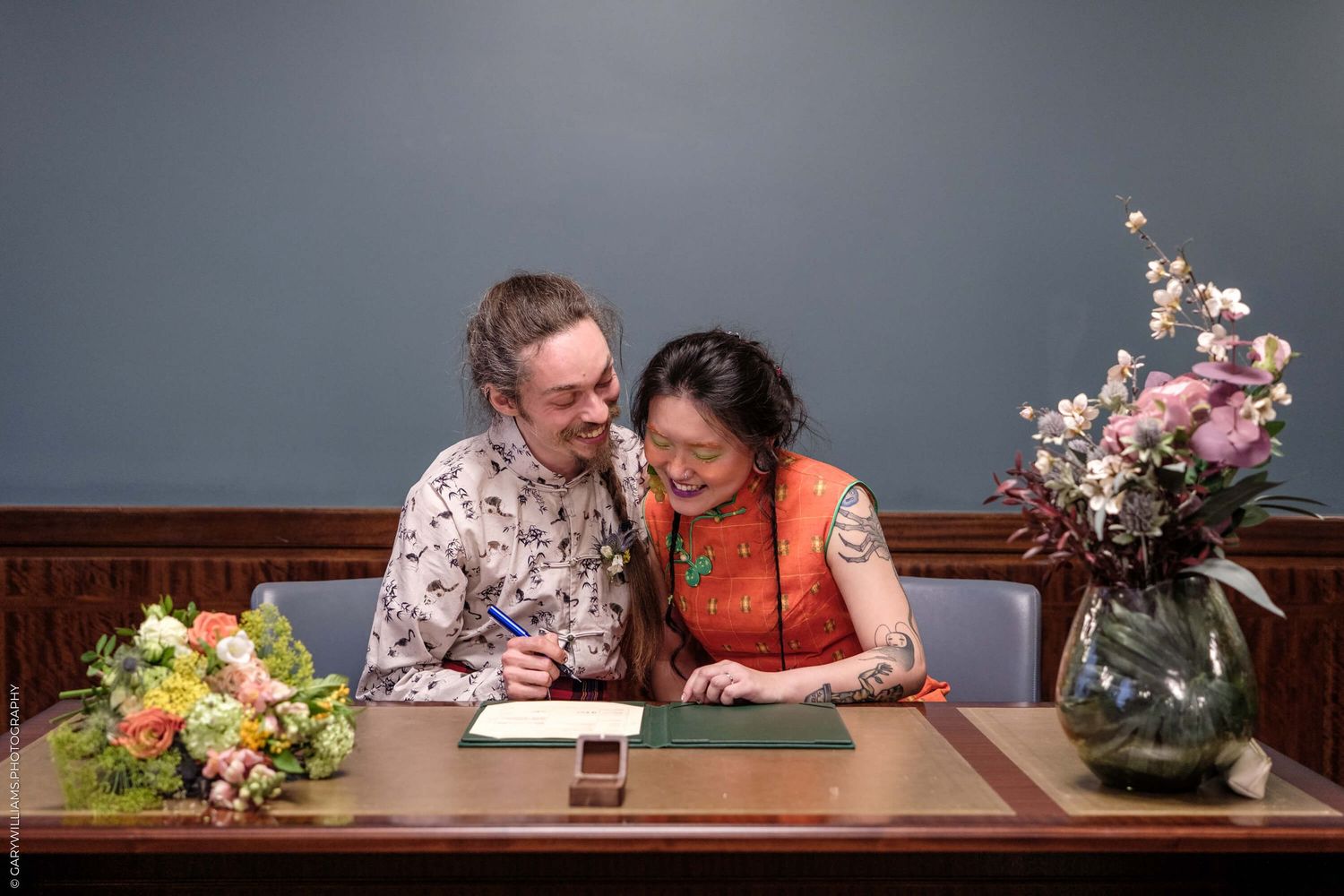 A couple shares a joyful moment while signing papers at a desk decorated with floral arrangements.
