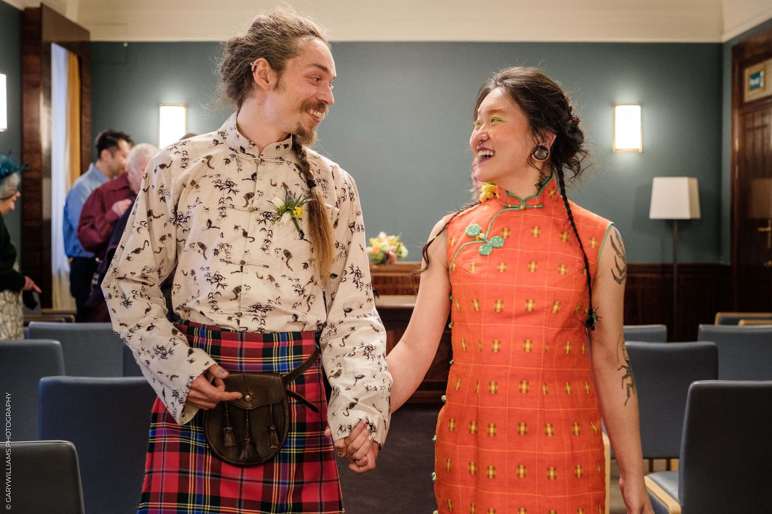 A person wearing a patterned shirt and kilt stands next to someone in an orange dress, both laughing together at an event.
