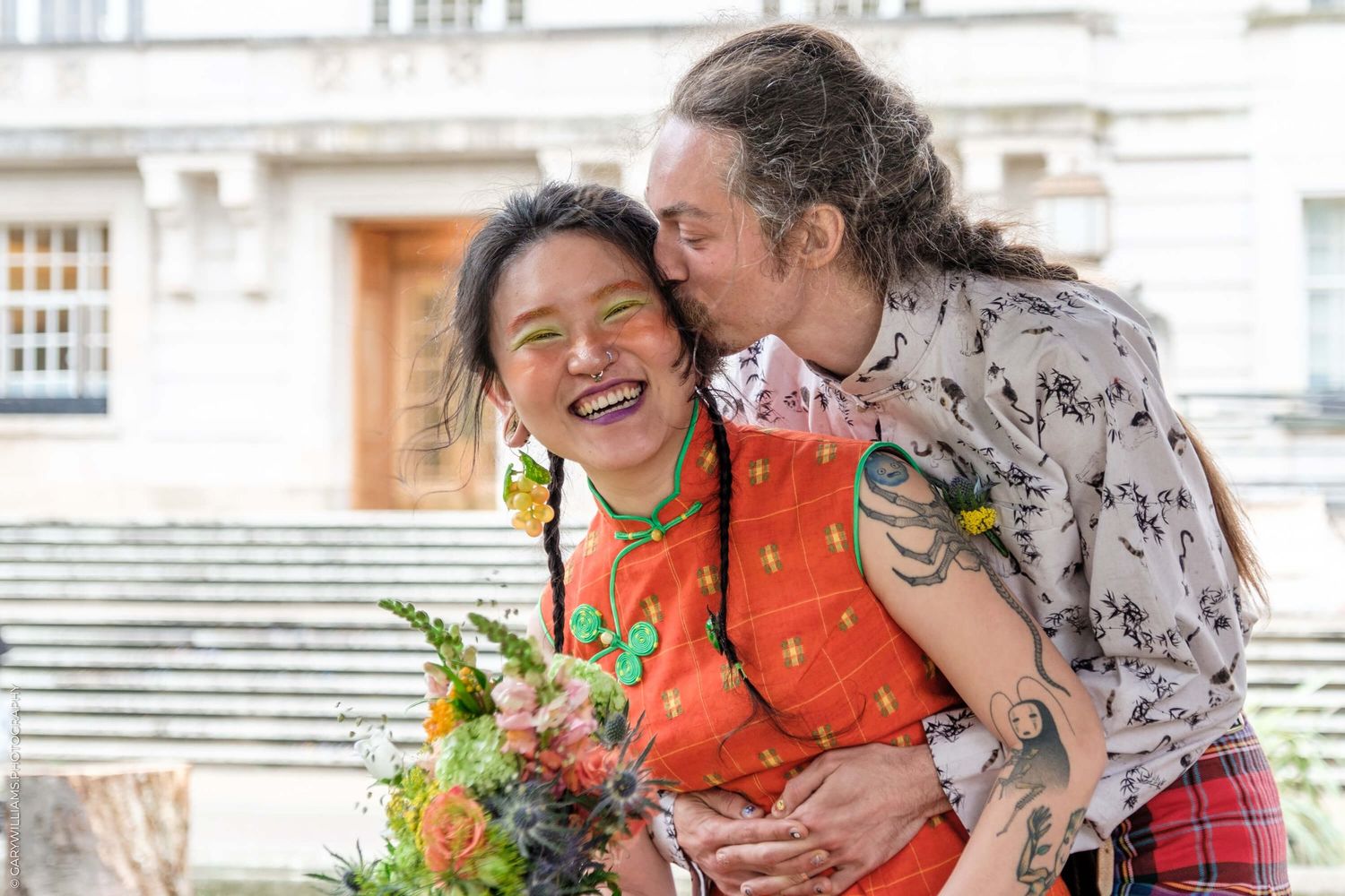 Two people share a joyful moment with laughter while holding a colorful bouquet of flowers outdoors.