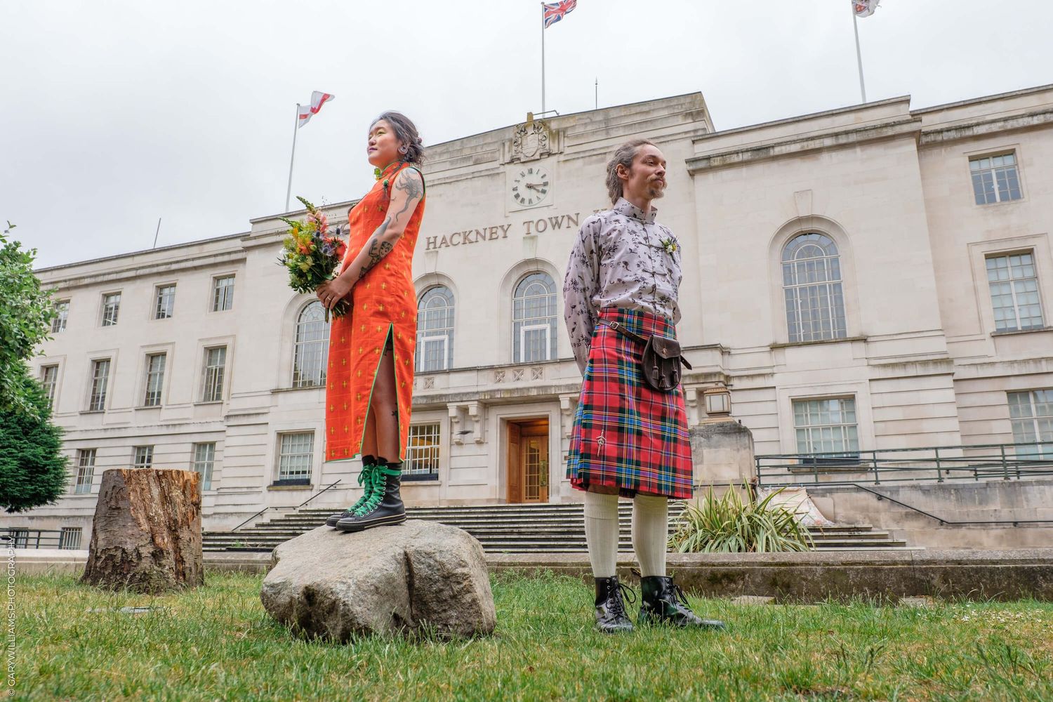 Two people stand outdoors wearing colorful outfits in front of a stately government building with flags on top.