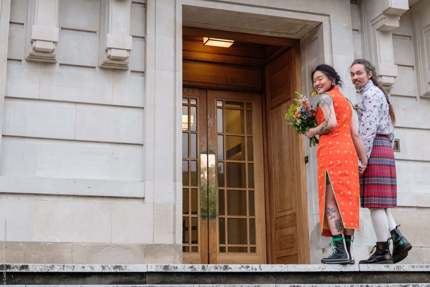 Two people stand in a doorway wearing colorful outfits - one in a bright orange dress and one in a plaid kilt.