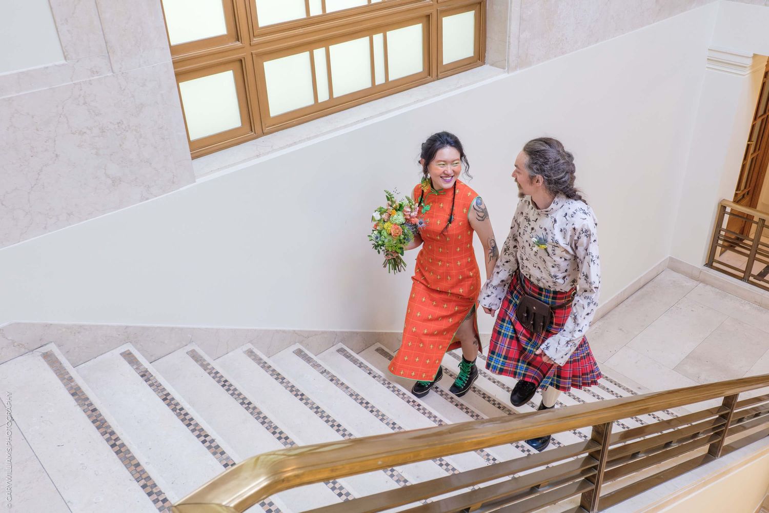Two people in colorful traditional outfits smile while walking up elegant white stairs with wooden railings near windows.