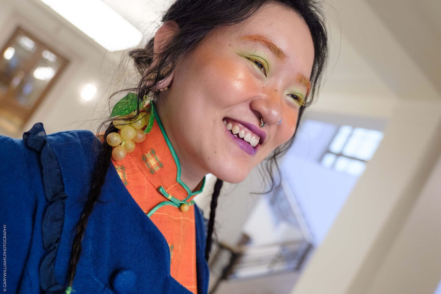 A person wearing blue clothing and orange scarf with bright yellow-green eyeshadow smiles at the camera.