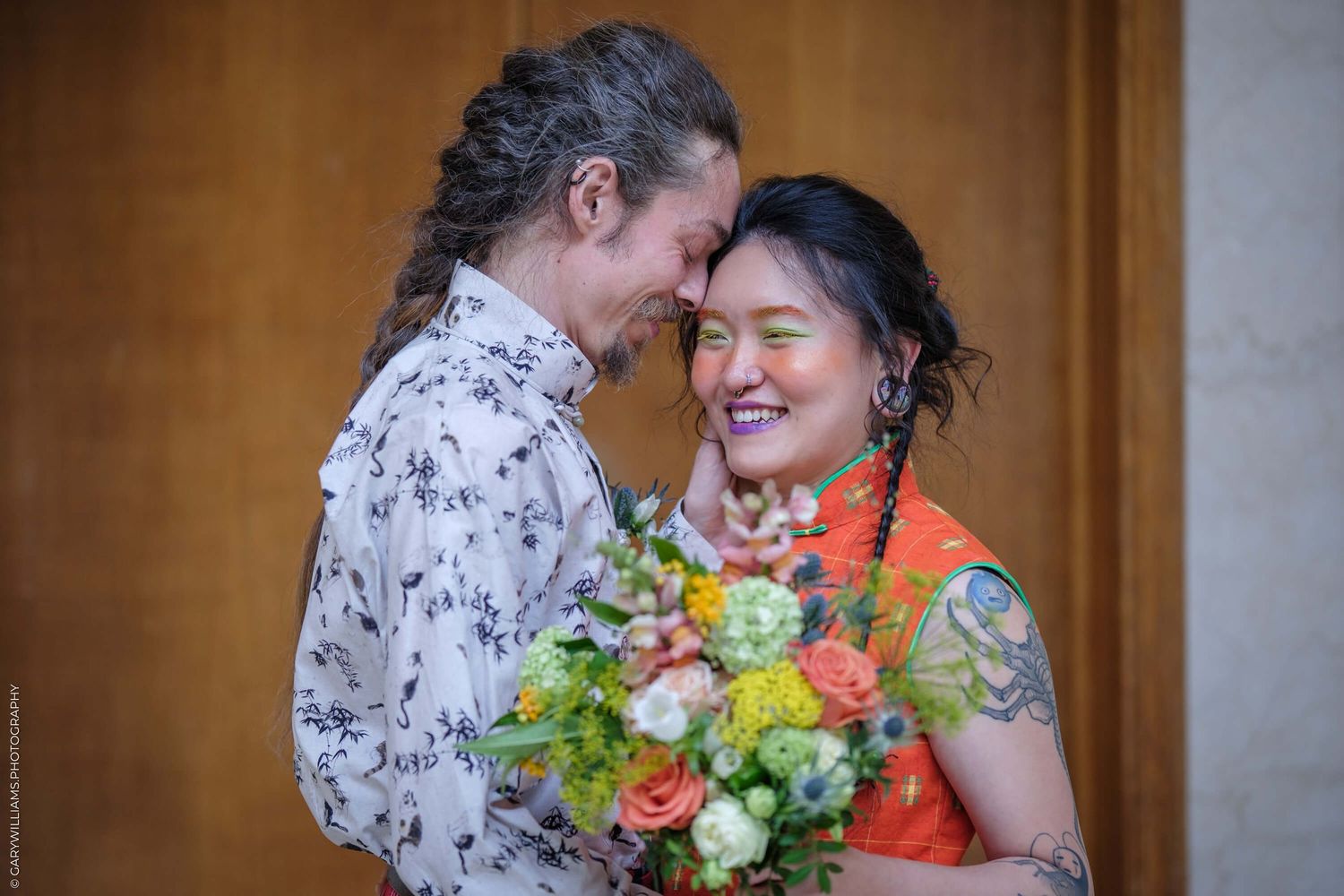 A couple shares a joyful intimate moment in front of a warm-toned background with orange and white floral decorations.