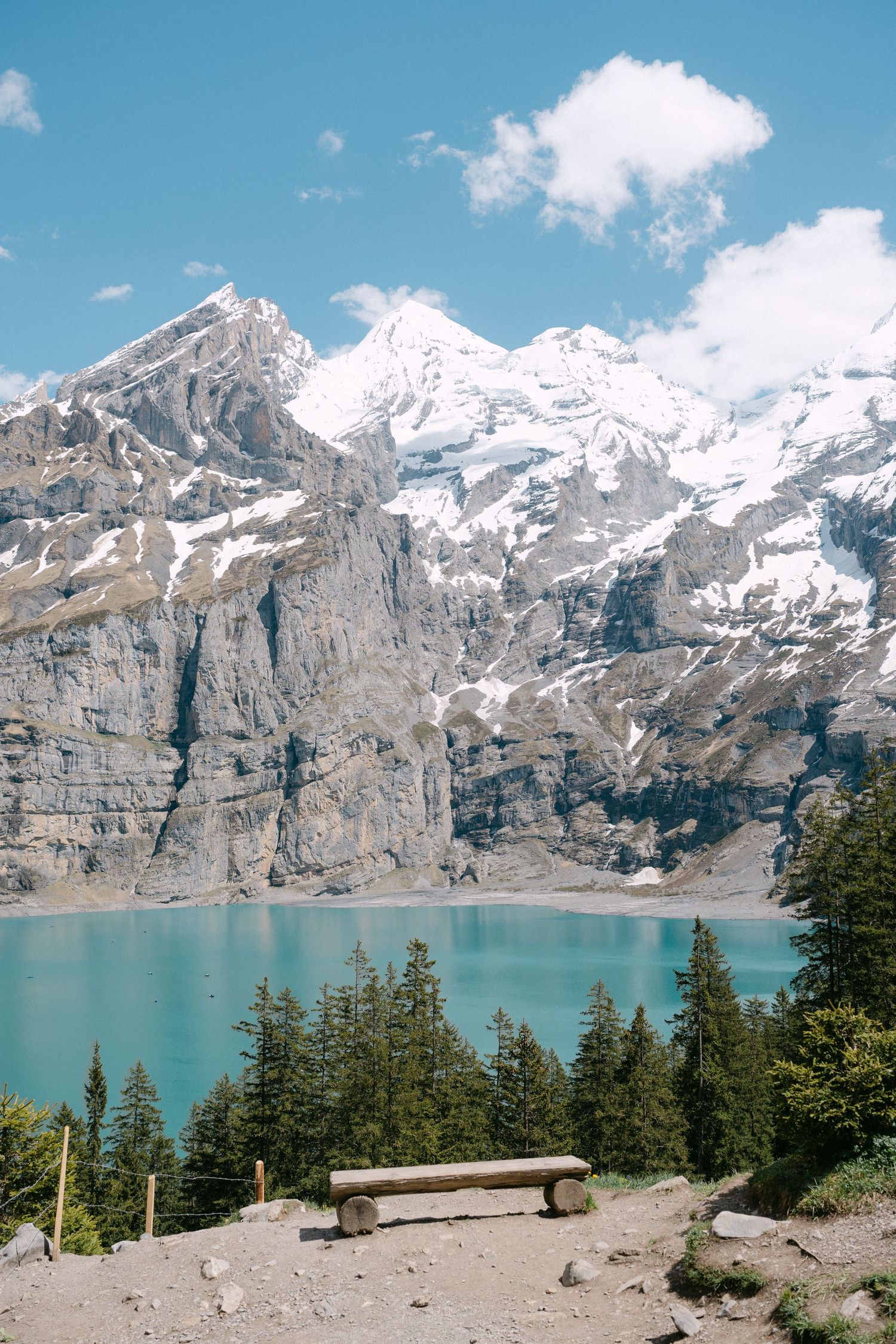 Rustic wooden bench overlooks turquoise alpine lake, Oeschinensee, with snow-capped mountains and evergreen trees in Swiss Alps landscape.