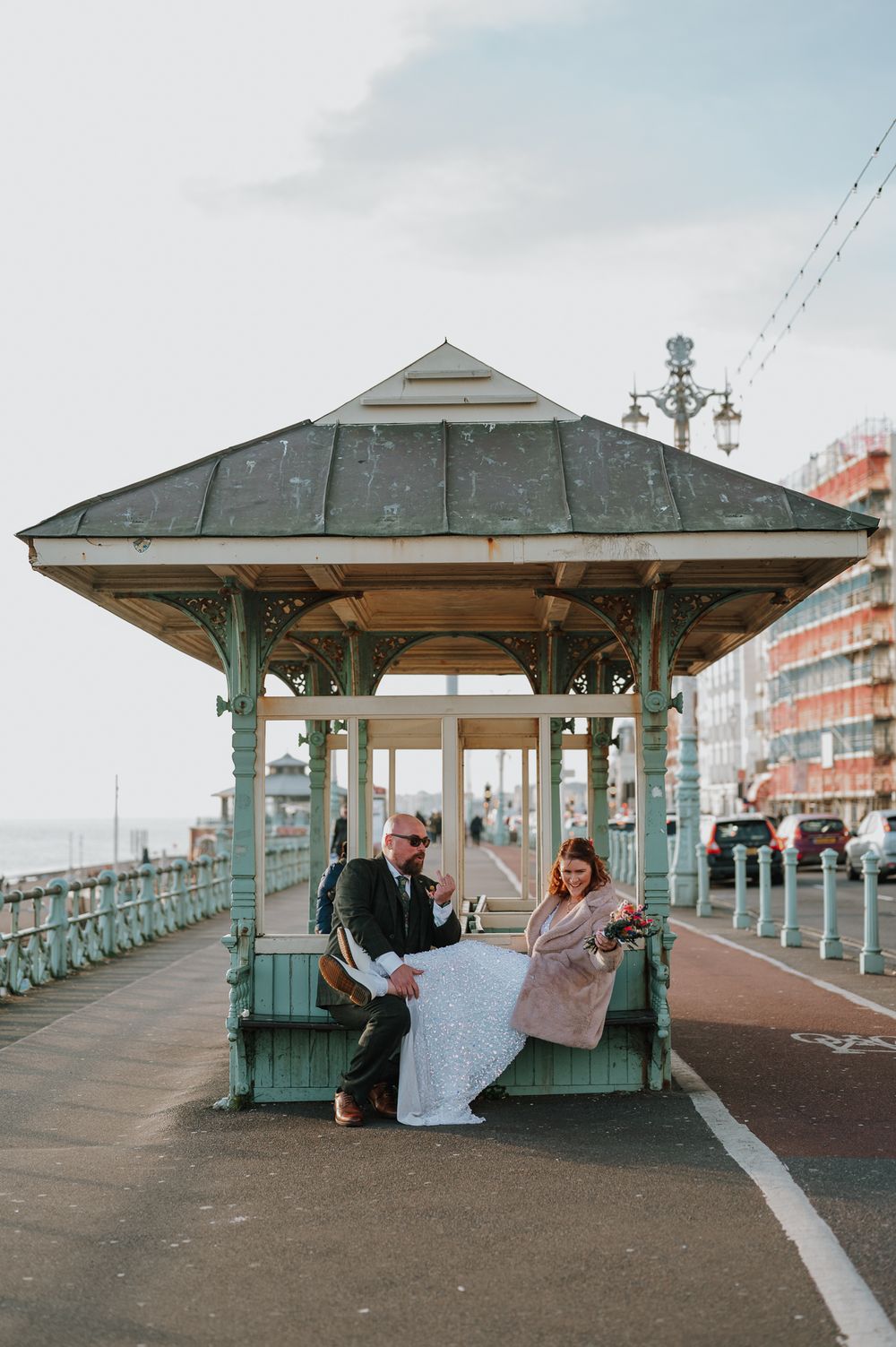 Two people sit together inside a seaside shelter on a promenade looking at what appears to be a map or document.