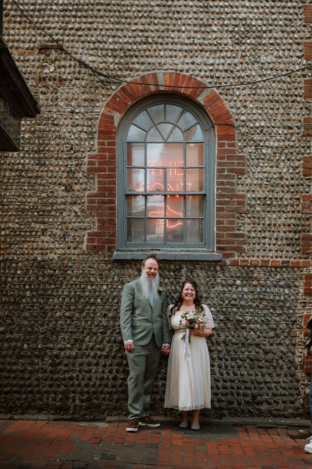 A couple stands in front of a vintage brick wall with an arched window on their wedding day.
