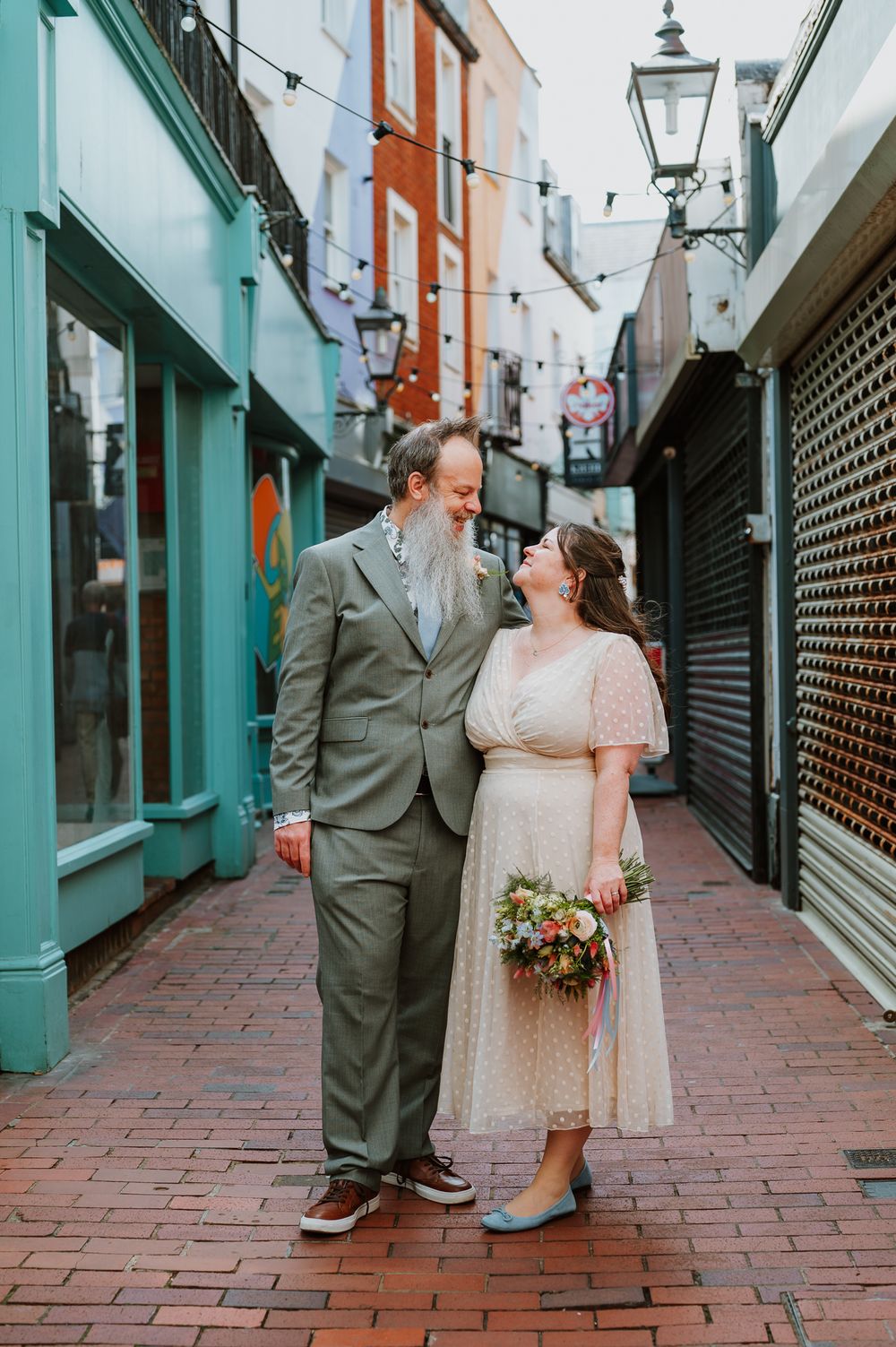 A couple stands close together on a charming brick alley with colorful buildings and string lights overhead in Brighton UK.
