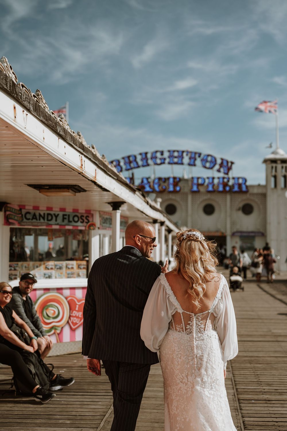 Newlyweds walking on Brighton Pier boardwalk past candy floss shop with iconic signage visible against moody sky.