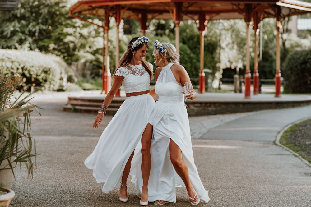 Two brides in elegant white wedding dresses dance together joyfully on a path under a red gazebo on their special day.