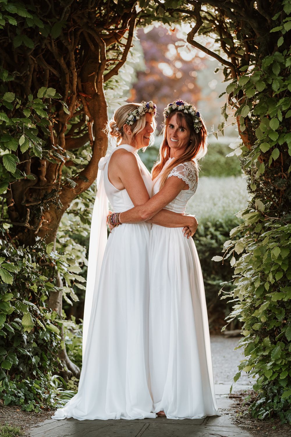 Two people in white dresses embrace in an outdoor garden setting framed by lush green vines.