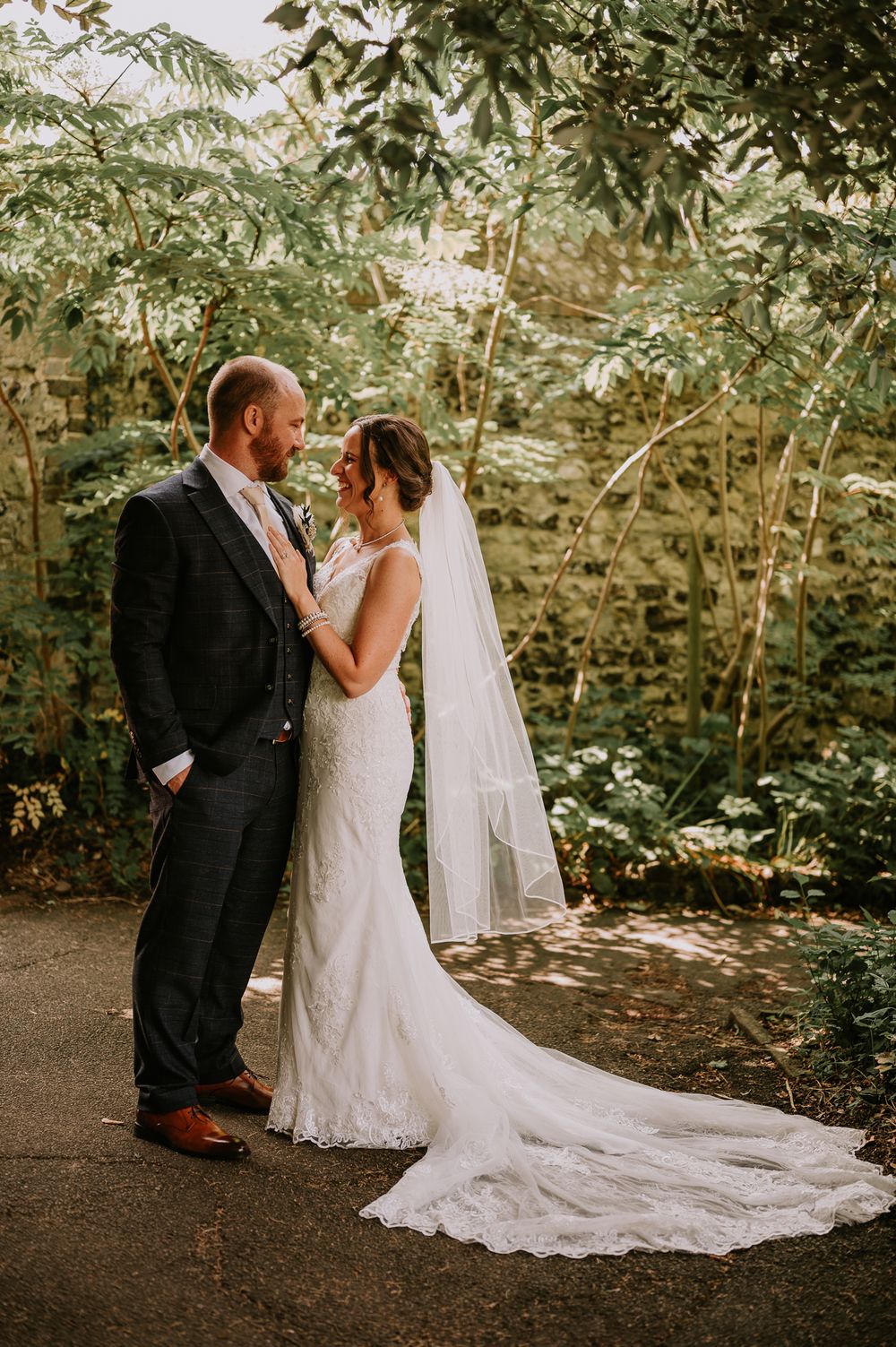 A romantic wedding couple embraces on a garden path surrounded by lush greenery and dappled sunlight.