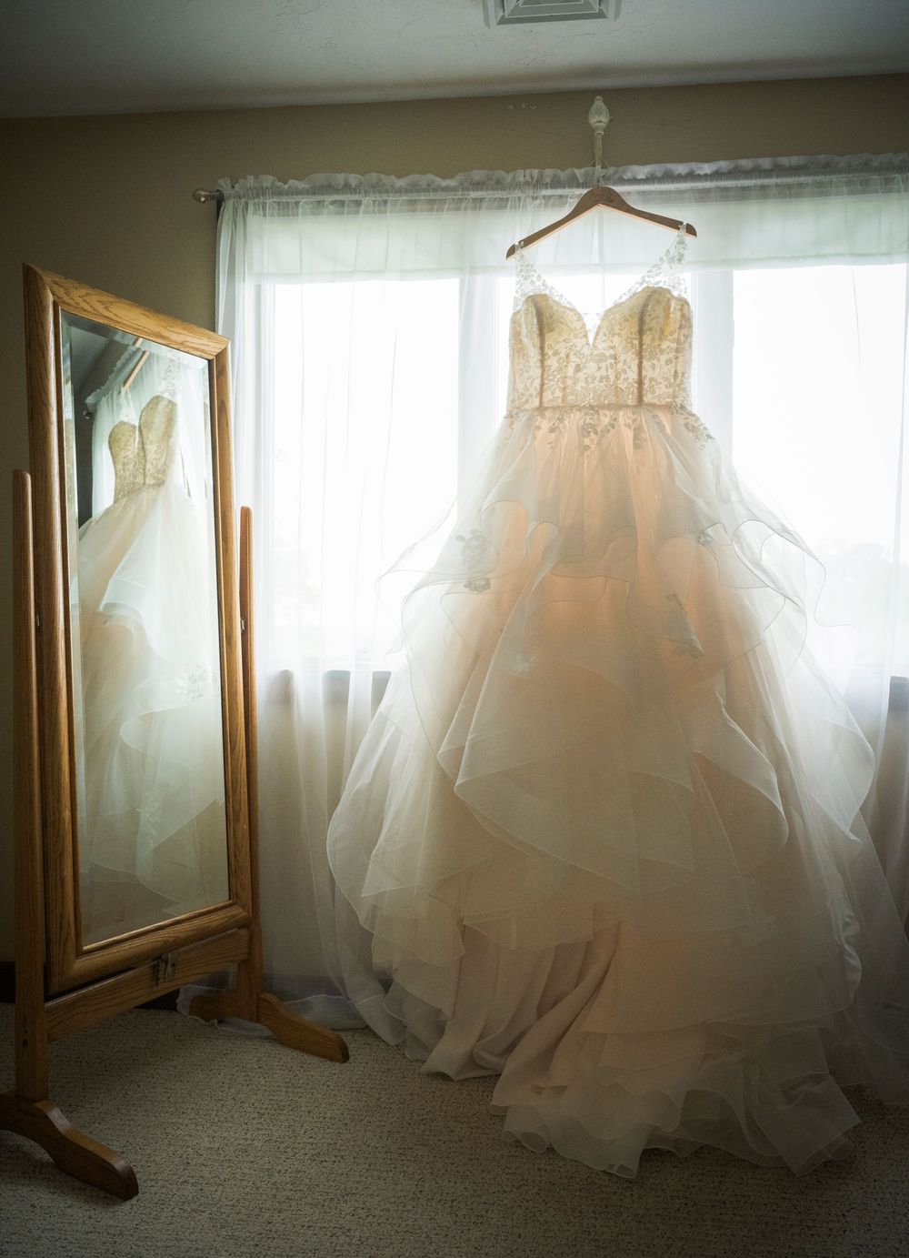 Elegant white wedding dress with layered tulle skirt hangs in front of window next to full-length mirror.
