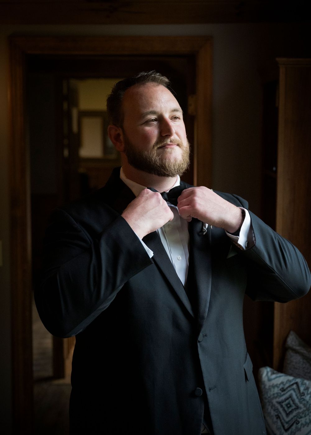 A person adjusts their bowtie while wearing a formal black tuxedo in a dimly lit room.