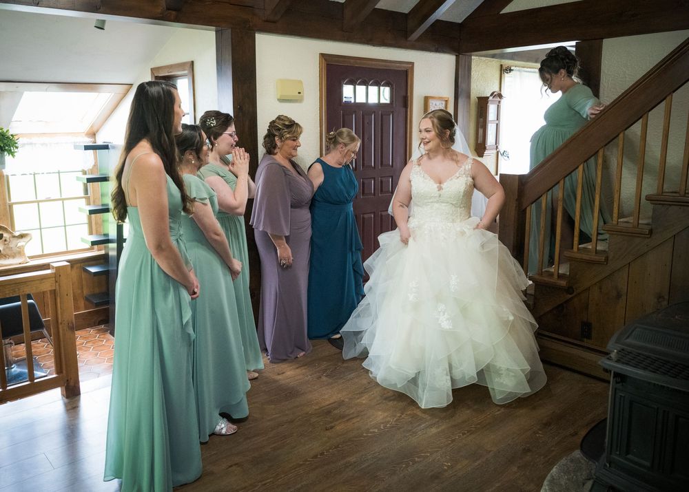 Bridesmaids in mint green and purple dresses admire the bride in her wedding gown by a wooden staircase.
