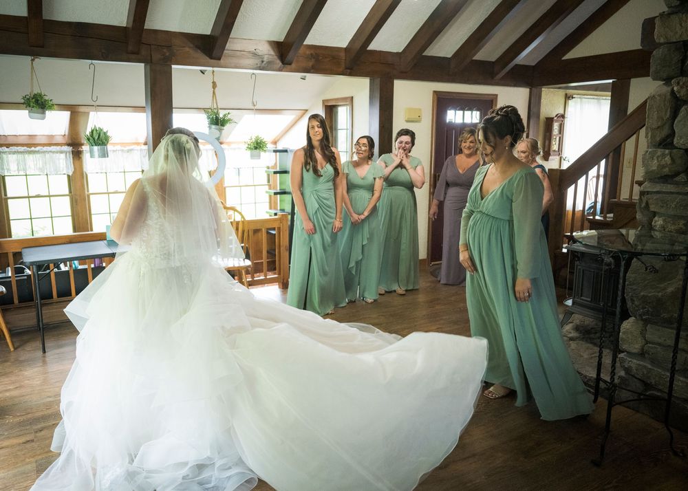 Bridesmaids in mint green dresses admire bride in white gown with long train in rustic indoor setting.
