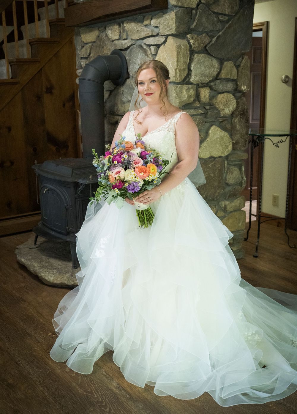 Bride in a flowing white ball gown poses with colorful floral bouquet against rustic stone wall interior.