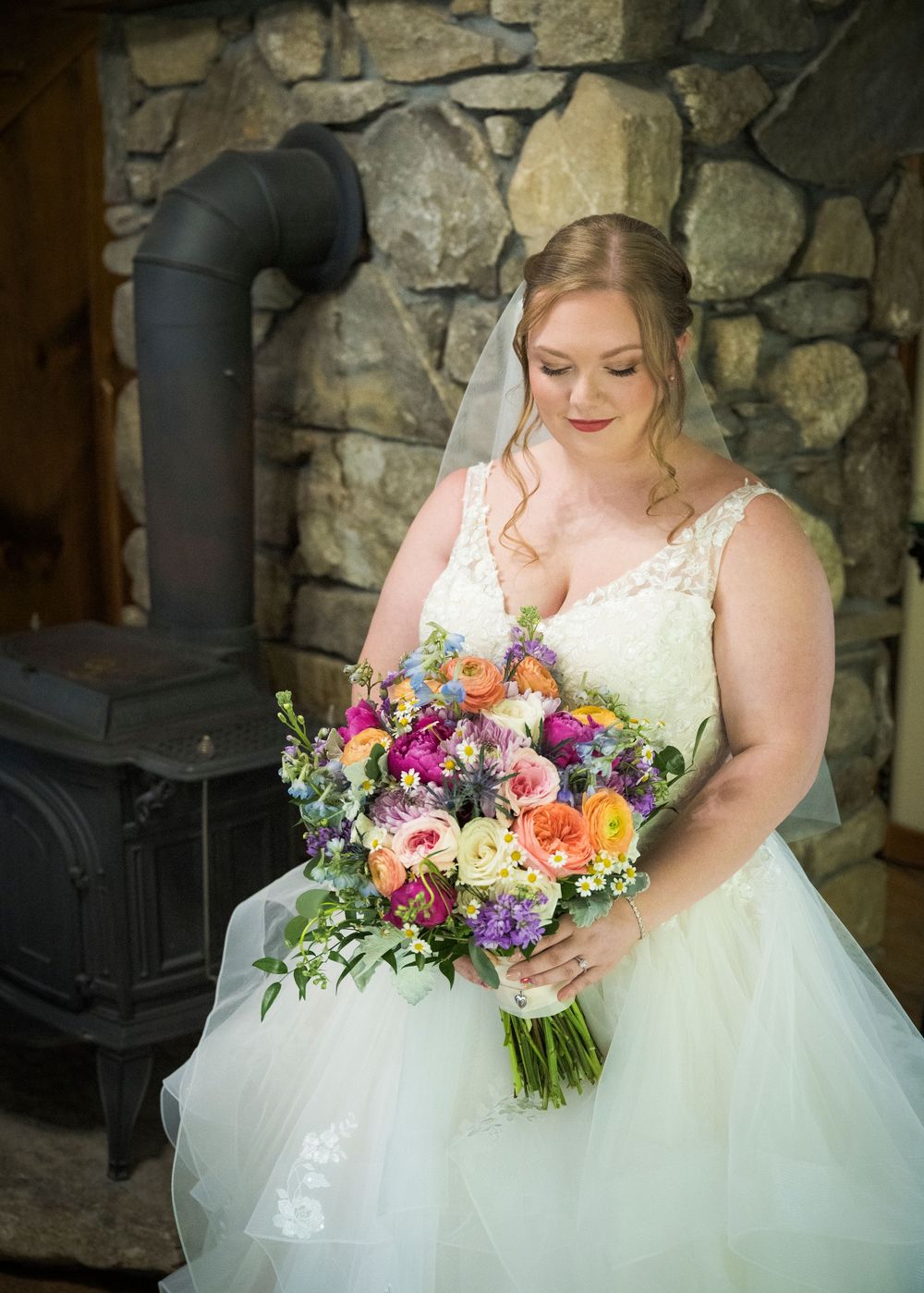 Bride in lace white dress holding vibrant multicolored wedding bouquet next to stone wall and wood stove.