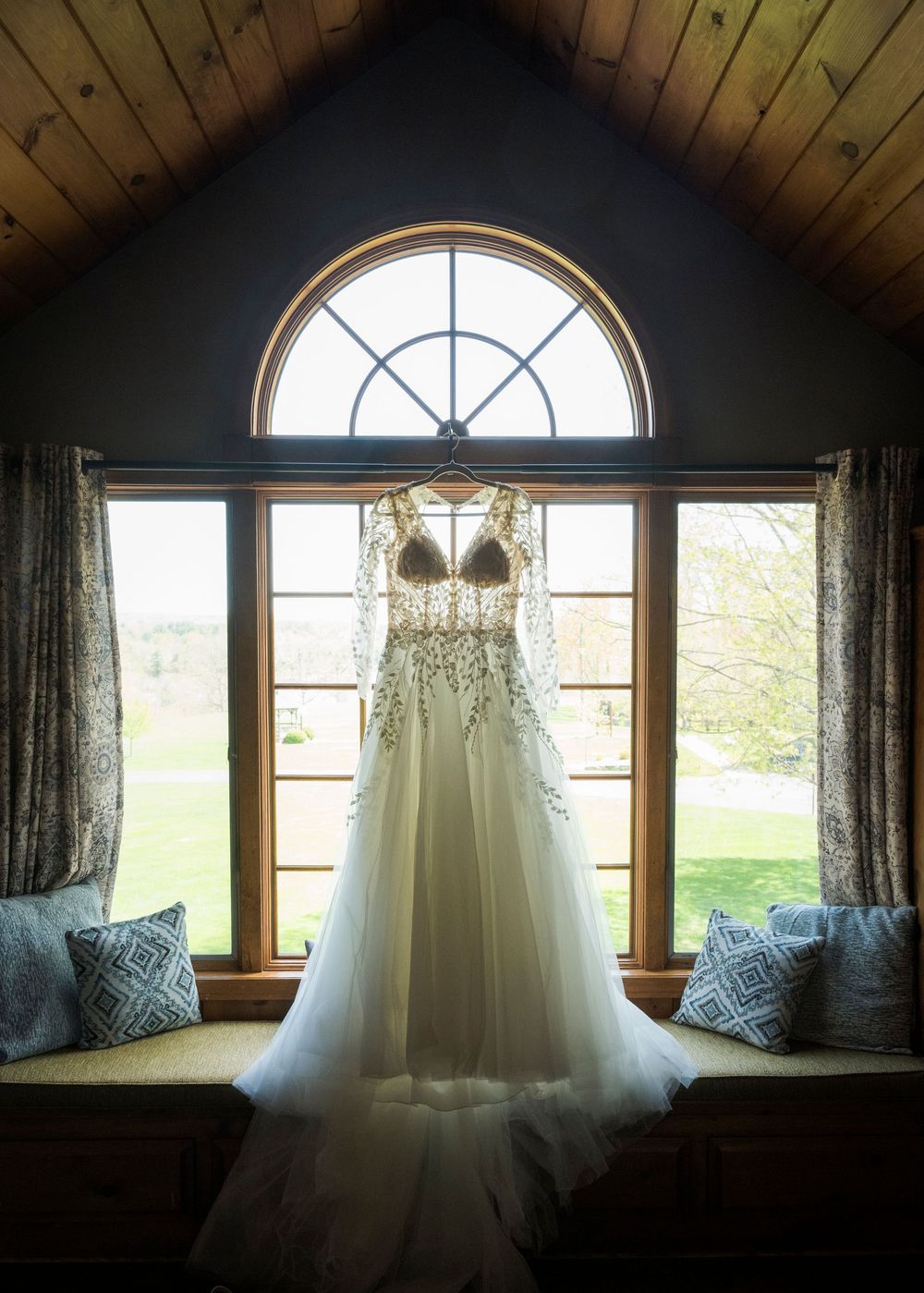 A white wedding dress hangs in front of an arched window with draped curtains and blue throw pillows on a window seat.