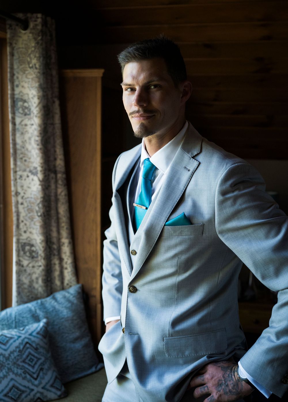 A formal portrait of someone wearing a light gray suit with turquoise tie in a rustic indoor setting.