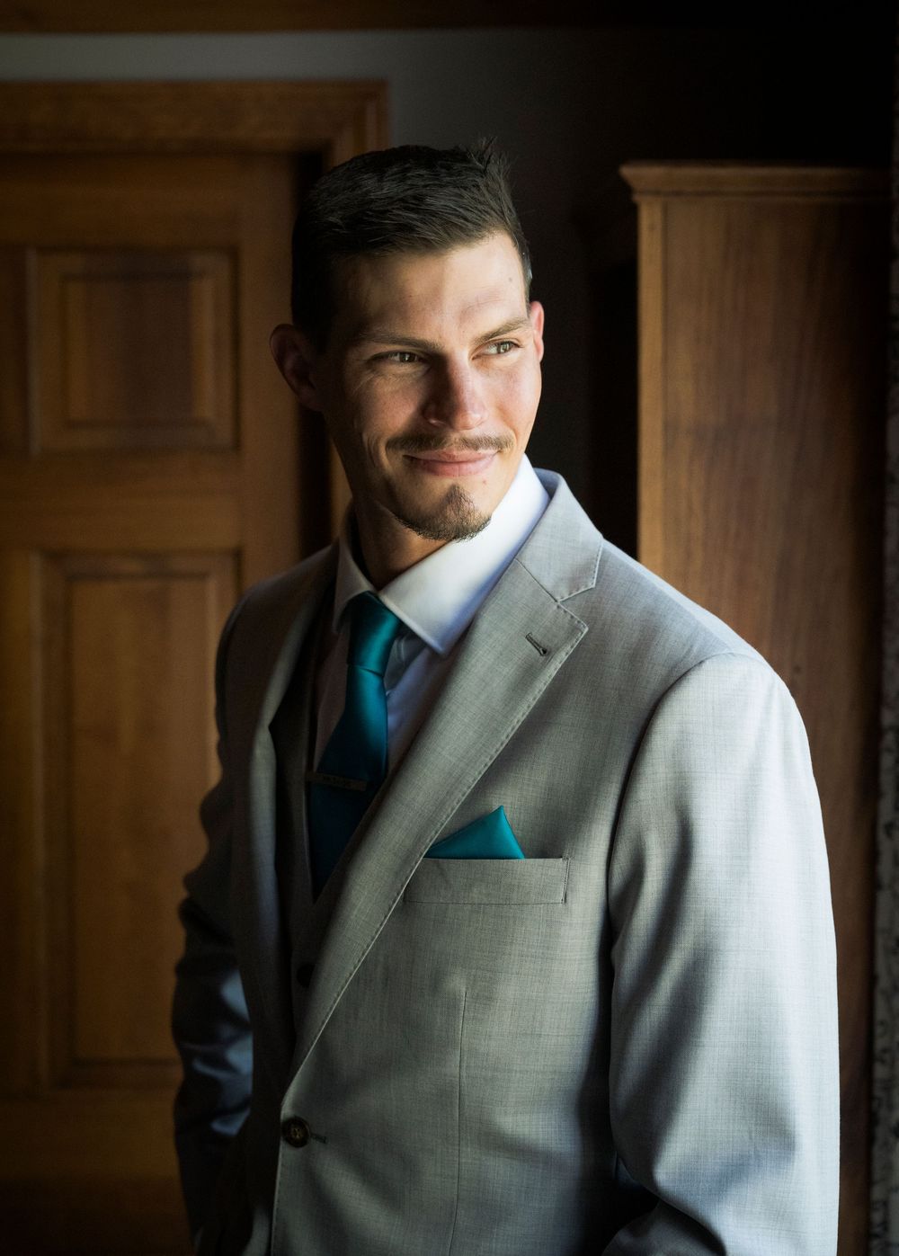 Person wearing a grey suit with teal tie and pocket square poses against wooden panel background.