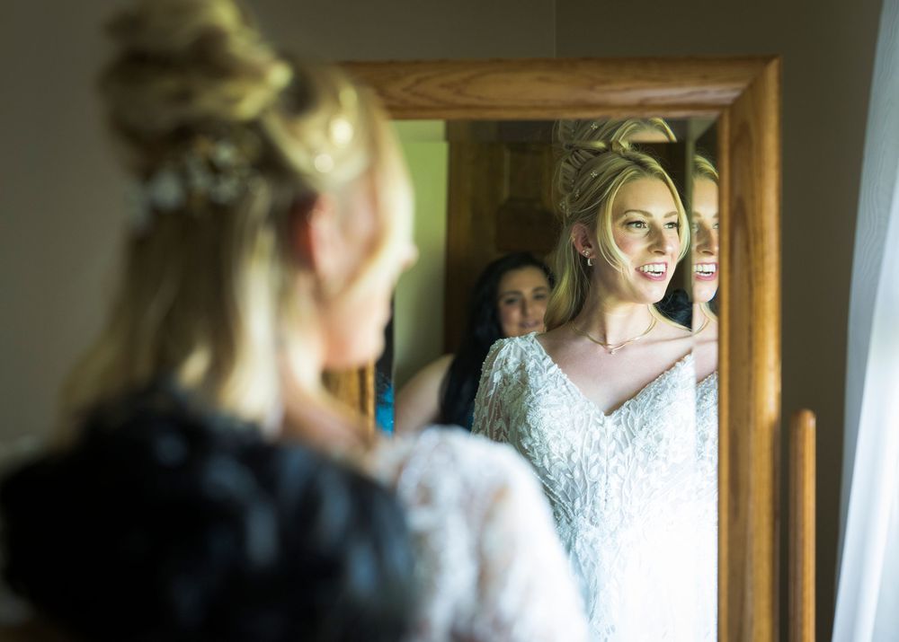 Bride looking in mirror while getting ready on wedding day with lace details visible.