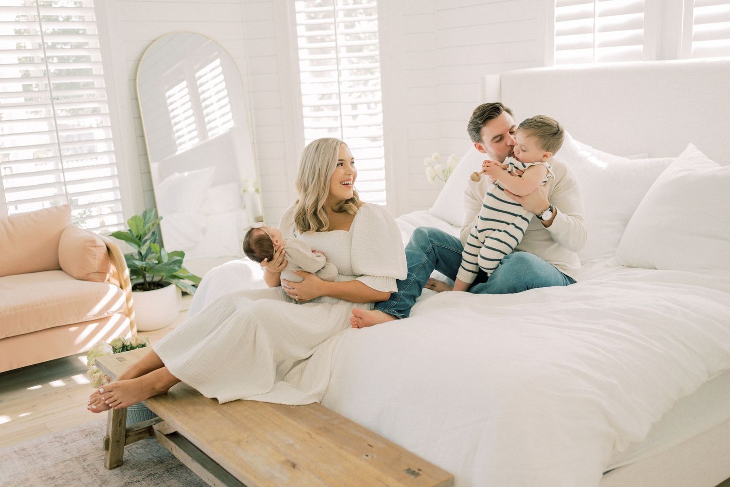 Family relaxes together on a white bed in a bright, sunlit bedroom with hardwood floors and white decor.