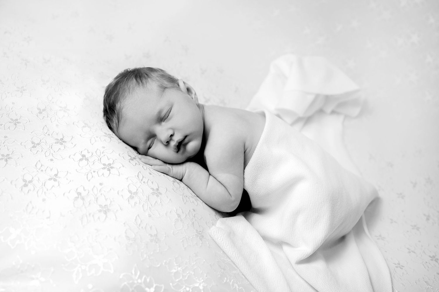 A sleeping newborn baby wrapped in white fabric rests peacefully against a soft textured white background.