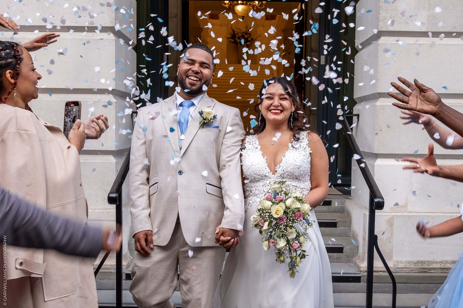 A joyful newlywed couple exits their wedding ceremony as guests shower them with white confetti on stone building steps.