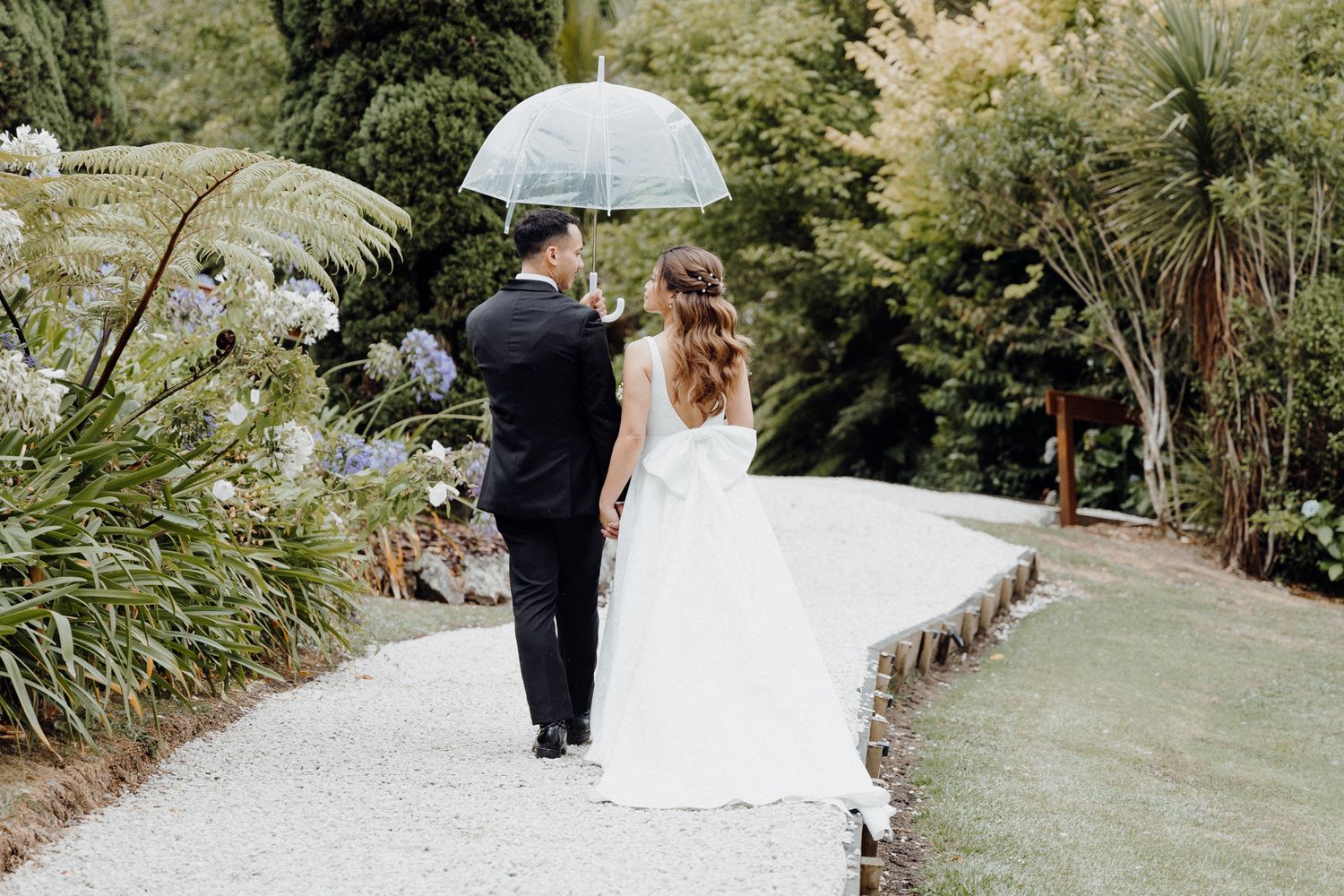 A couple walks along a garden path under a clear umbrella surrounded captured by Apertured Media Auckland Wedding Photographer.