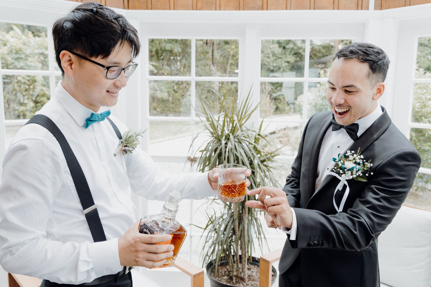 Two people in formal attire share drinks while standing in a bright sunlit room captured by Apertured Media Auckland Wedding Photographer.