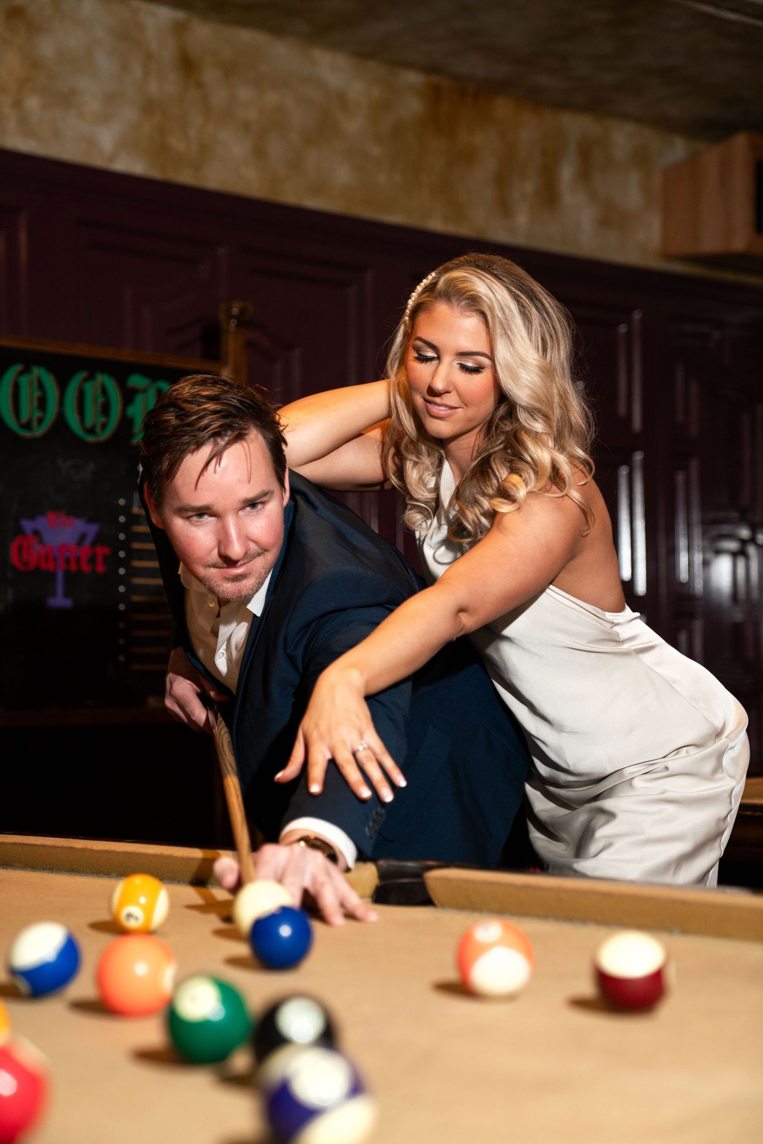 Playful moment at pool table in dimly lit bar with vintage decor. Engagement Session at The Lafayette Hotel in San Diego. JPPC
