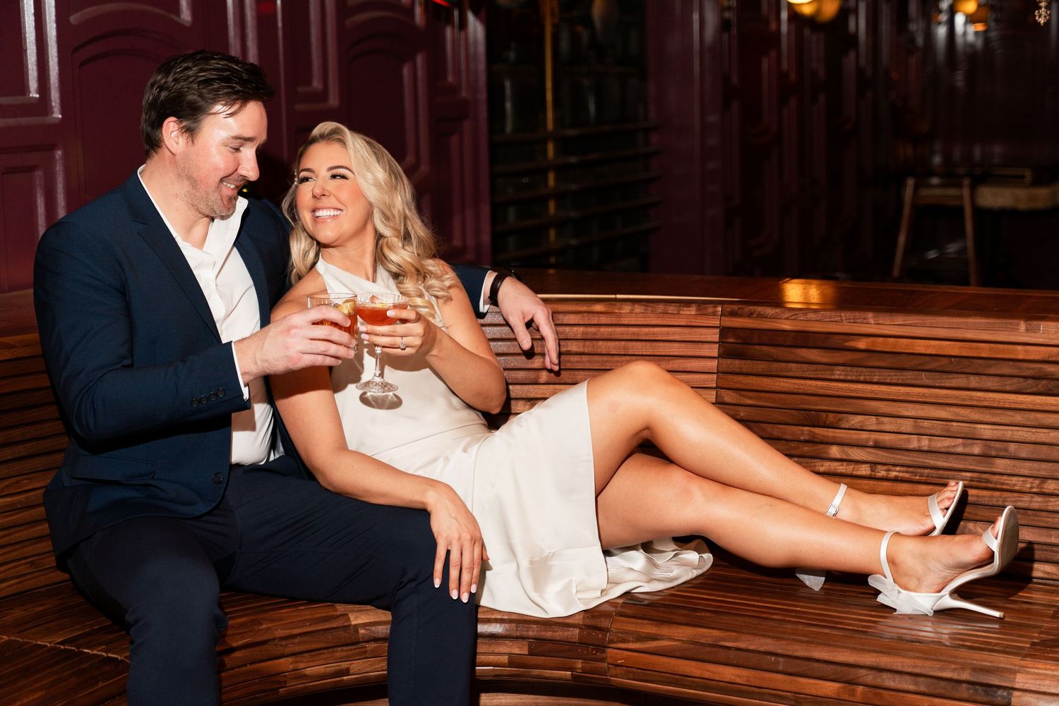 Couple shares drinks while relaxing on wooden bench in atmospheric bar setting. Engagement Session at The Lafayette Hotel in San Diego. JPPC