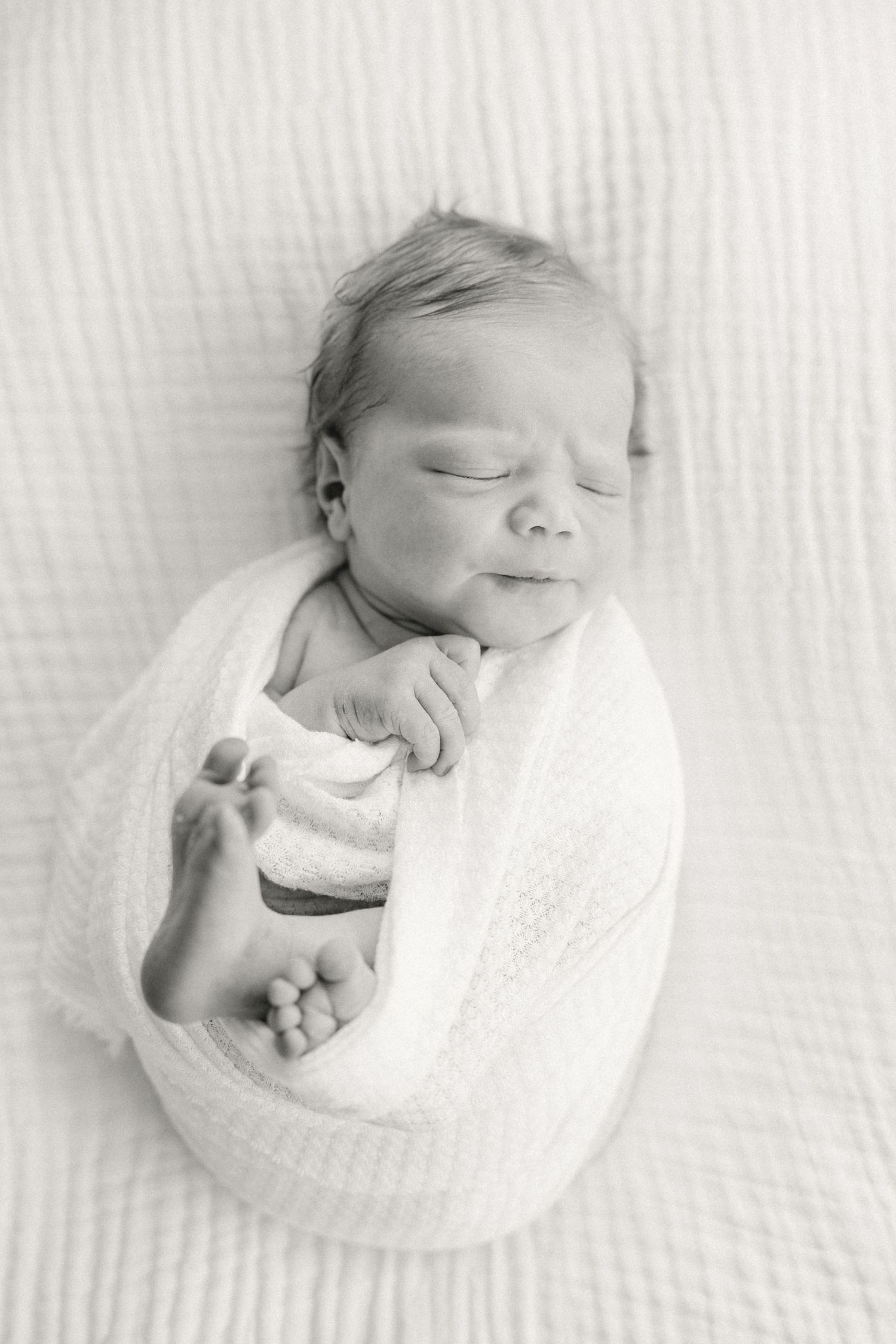 A sleeping newborn baby wrapped in a soft white blanket lies peacefully against a textured white background.