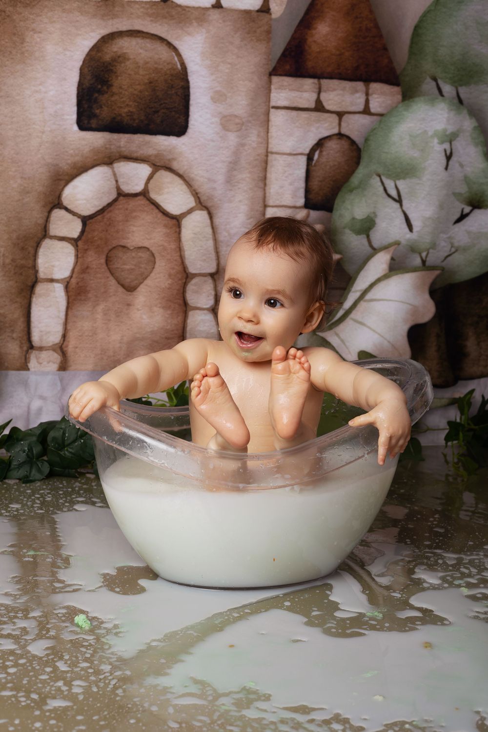 A small child sits joyfully in a clear glass mixing bowl against a whimsical fairytale backdrop with stone walls and trees.