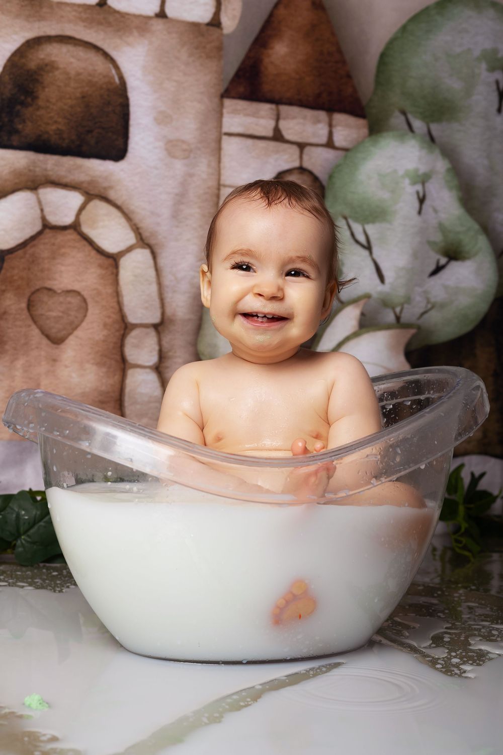 Baby sits in a clear glass bowl filled with milk against a whimsical castle backdrop with painted scenery.
