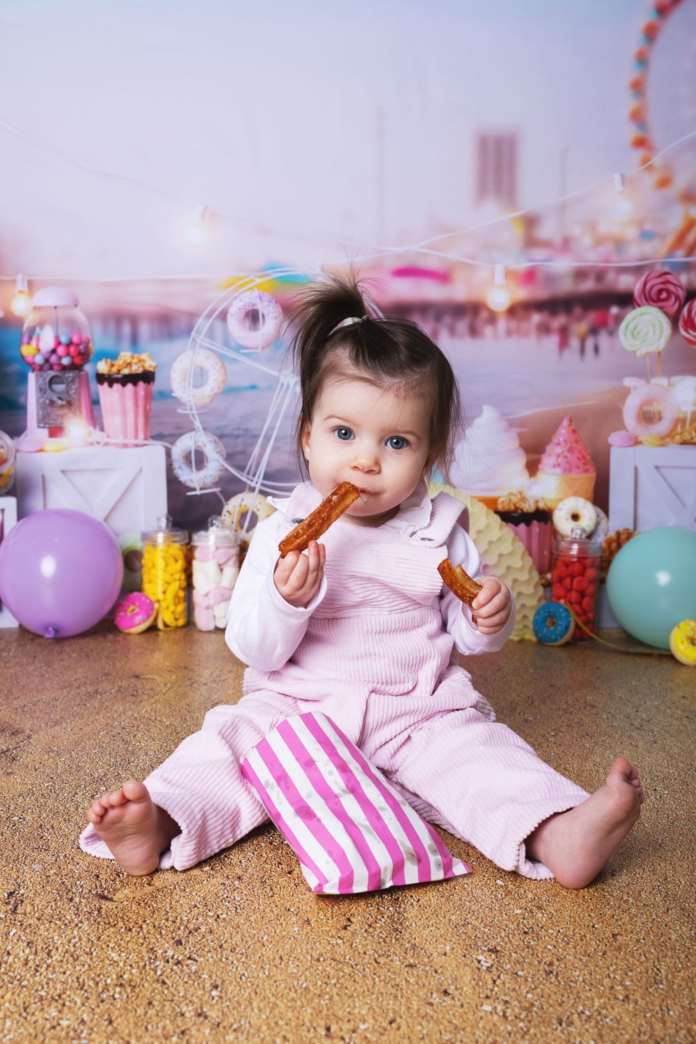 A baby in pink sleepwear enjoys a pretzel surrounded by colorful balloons and carnival-themed decorations.