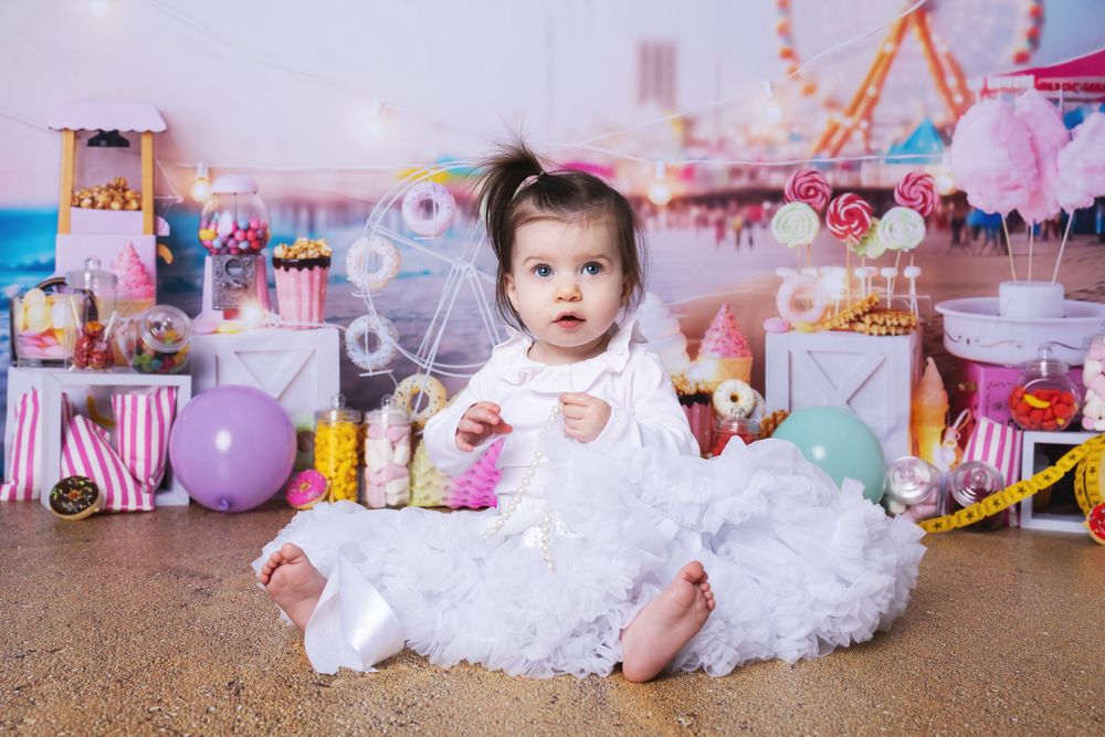 A baby wearing a fluffy white dress sits among carnival-themed decorations with pink and purple balloons and candy stripes.