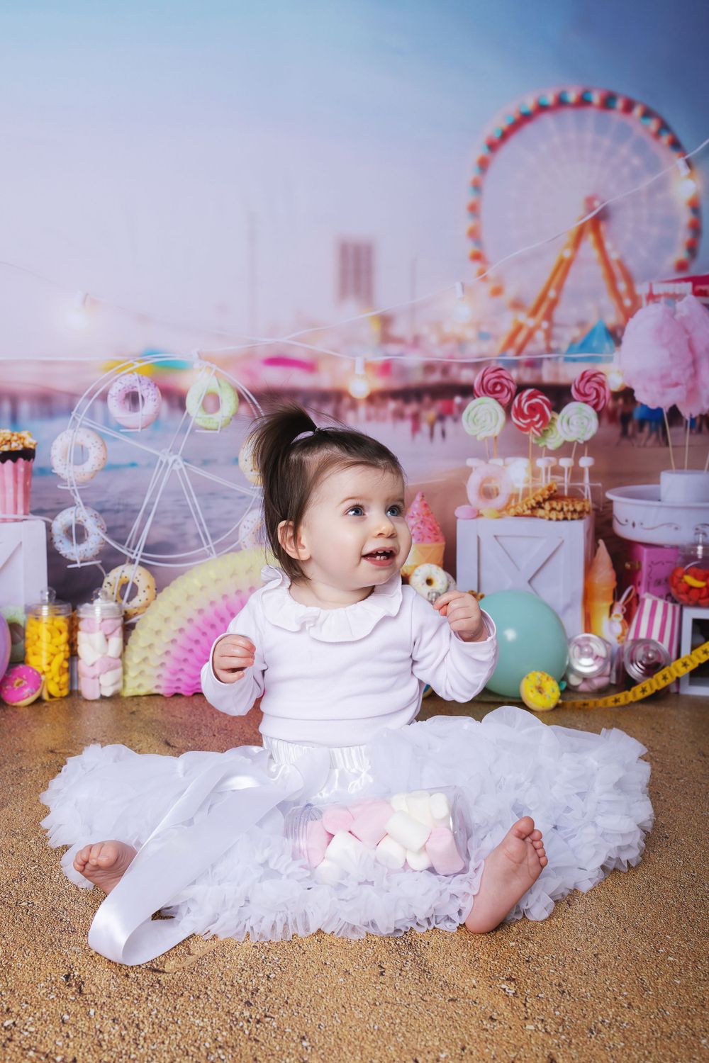Baby in white dress sitting against carnival backdrop with ferris wheel and amusement rides in dreamy pastel colors.