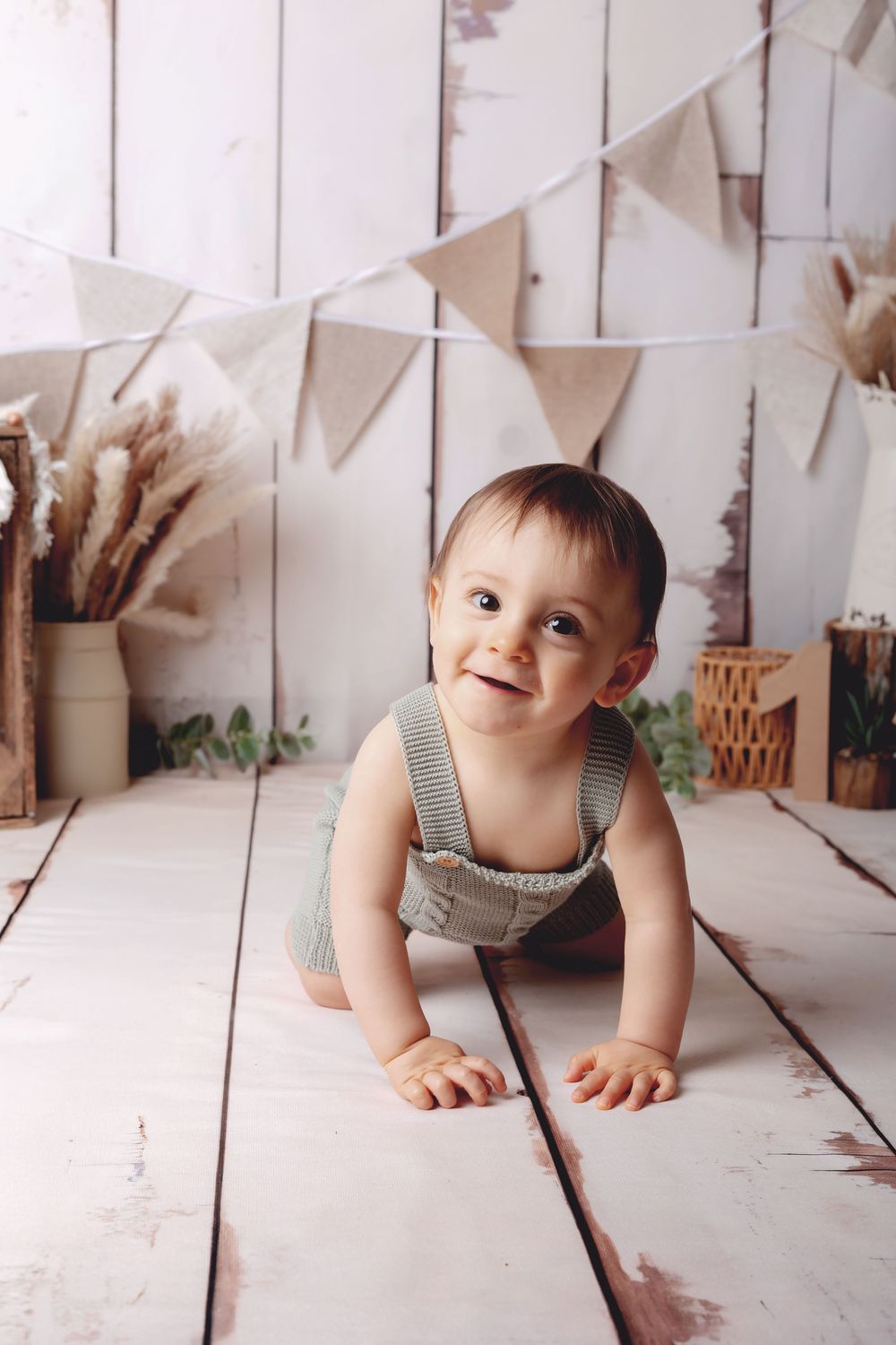 A baby in gray overalls crawls on white wooden floors with bunting flags and dried flowers decorating the background.