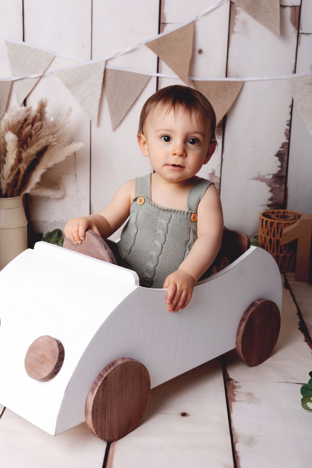 A small child wearing gray overalls sits in a white wooden toy car with brown wheels under neutral party decorations.