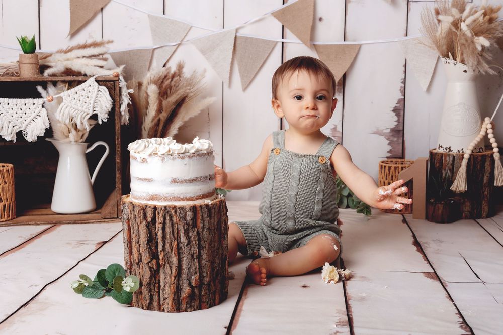 First birthday celebration setup with rustic wood decor, white bunting, pampas grass and a white cake on wooden stump.