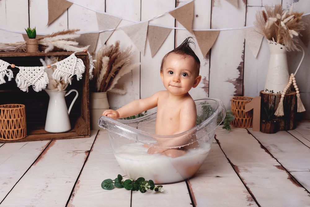 Baby sitting in a clear bathtub with milk bath surrounded by vintage decor and bunting flags on white wooden floor.