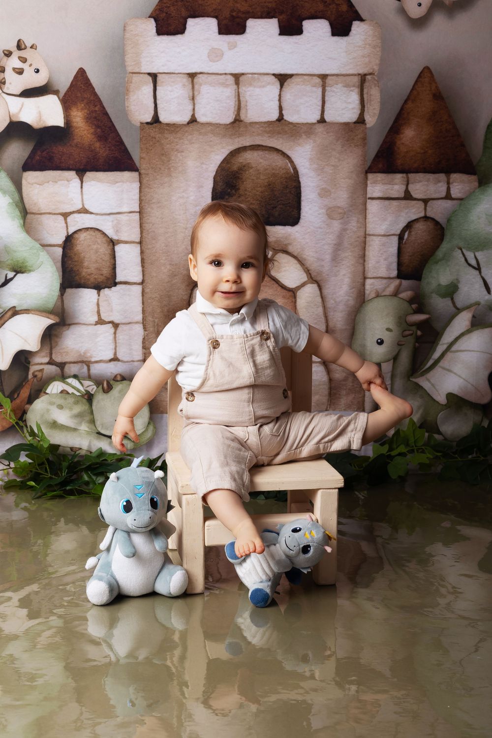 Adorable baby in beige overalls sits on bench with plush toys against fairy tale castle backdrop and greenery.
